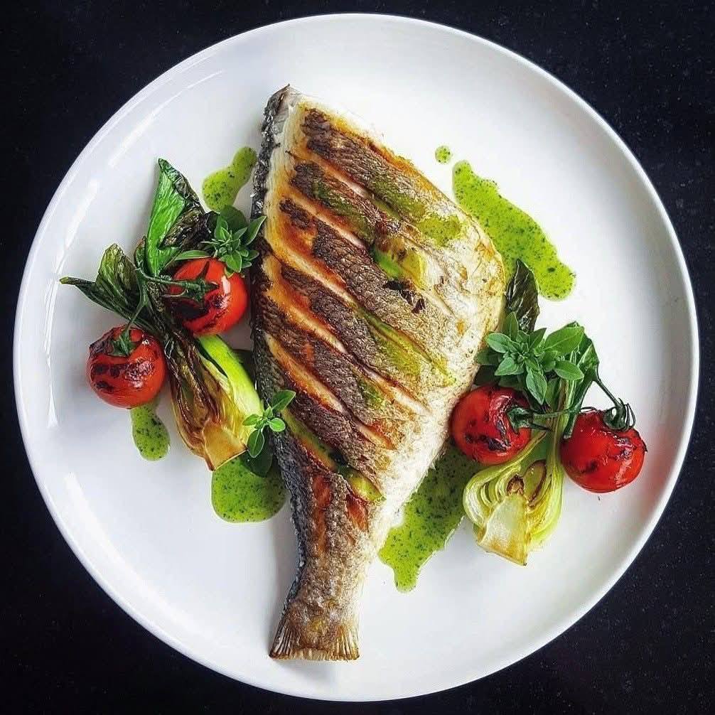 Grilled bass fillet on a white plate with green sauce, cherry tomatoes, and vegetables