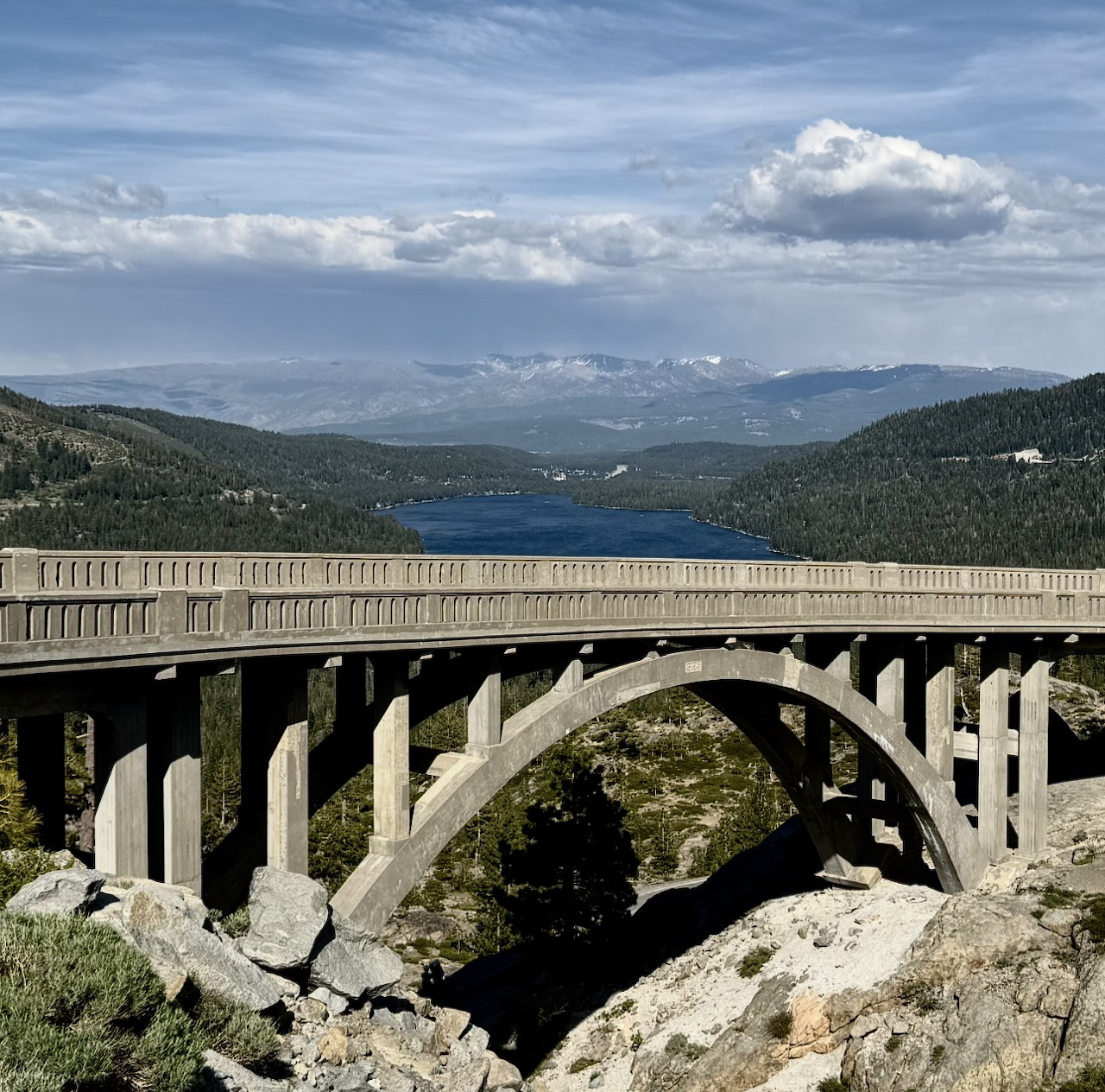 Concrete arch bridge over a rocky gorge with a lake, hills, and distant mountains beneath clouds.