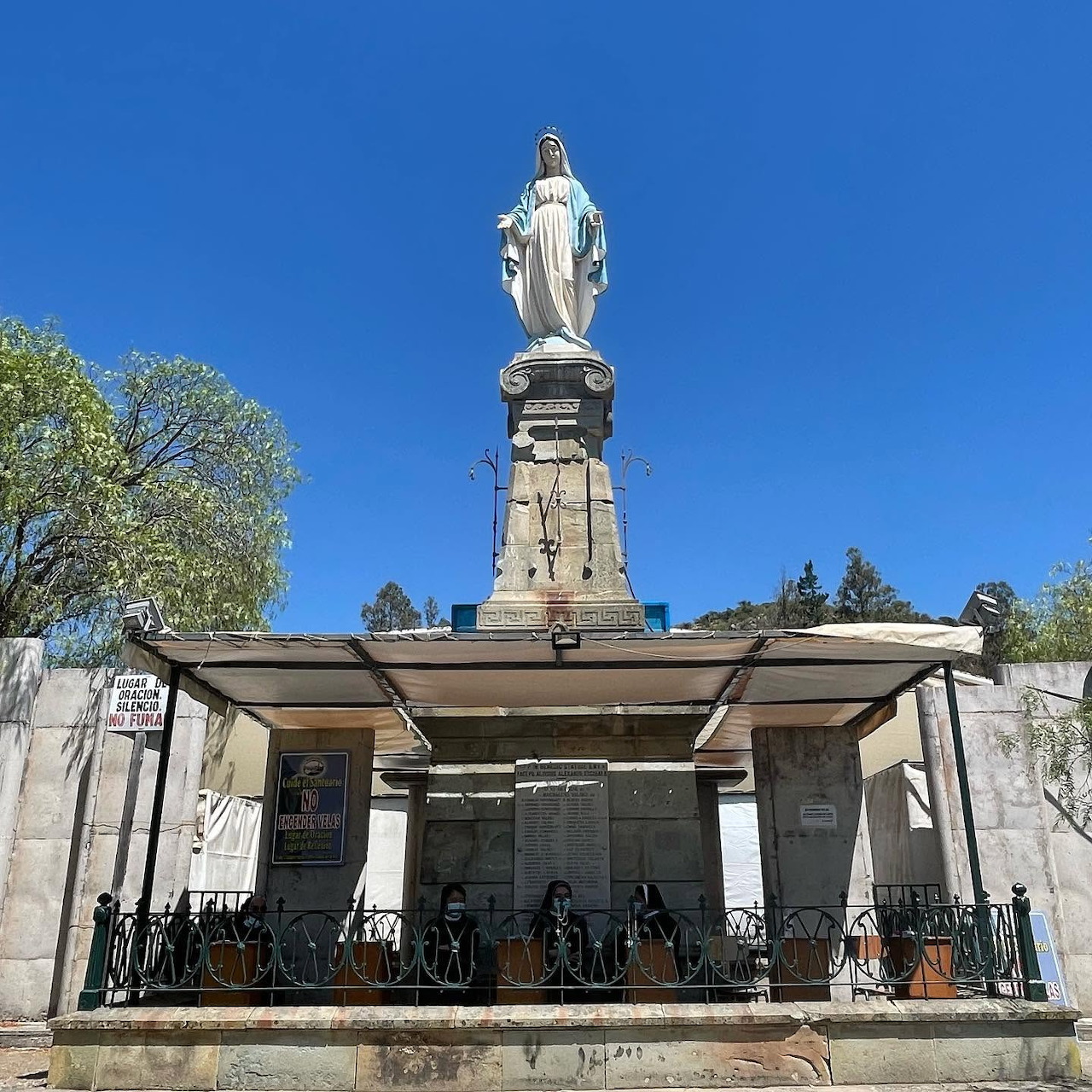 White Virgin Mary statue on tall monument with columned shelter beneath, blue sky background