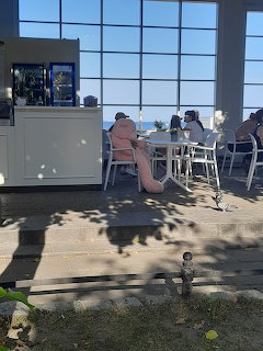 People relaxing at an outdoor cafe terrace with white furniture and large glass windows