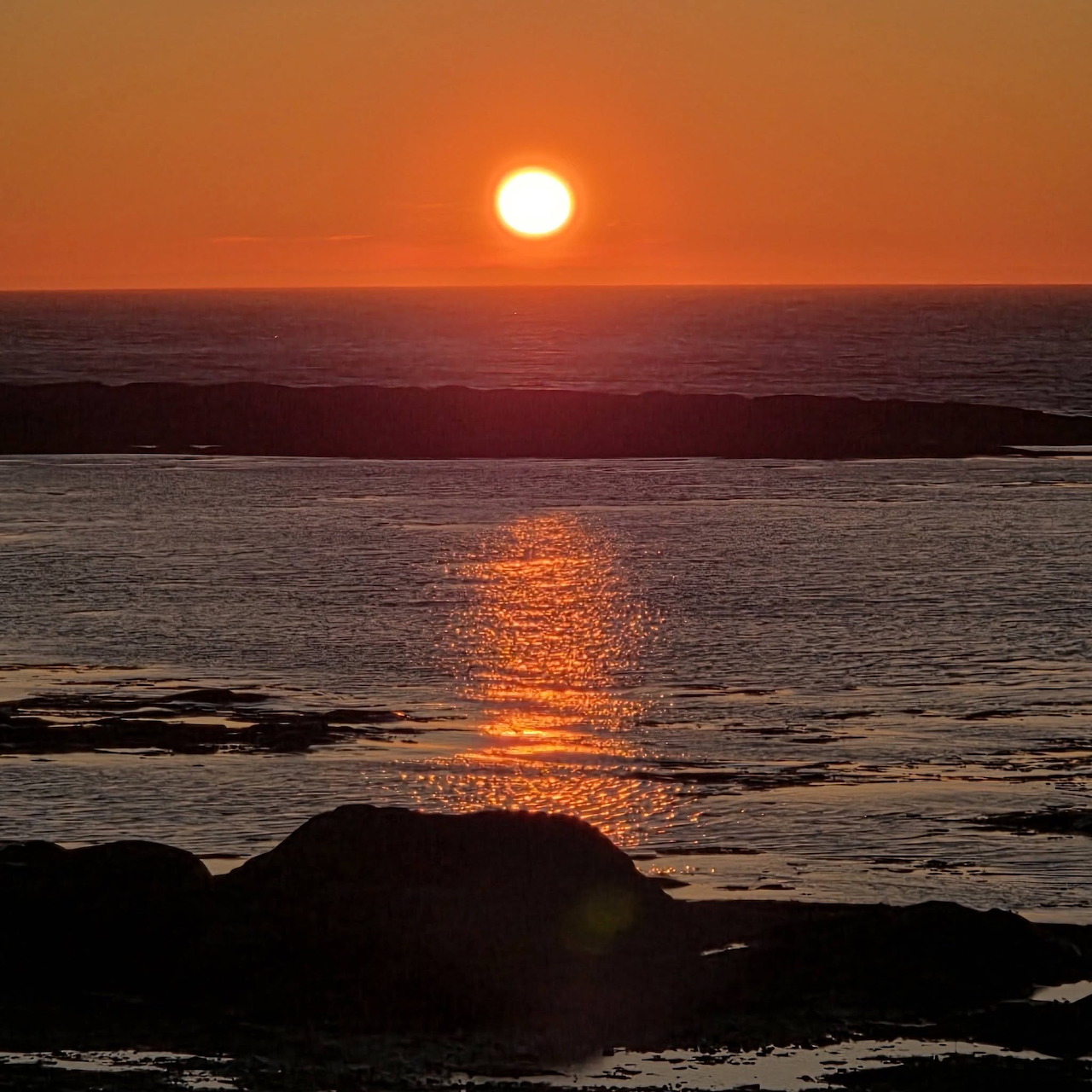 Orange sunset over the ocean with reflected light on calm water and dark rocks in the foreground