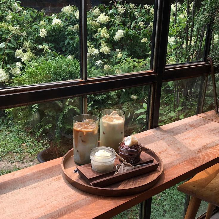 Two iced coffee drinks and a small dessert on a wooden counter by a window with garden greenery outside