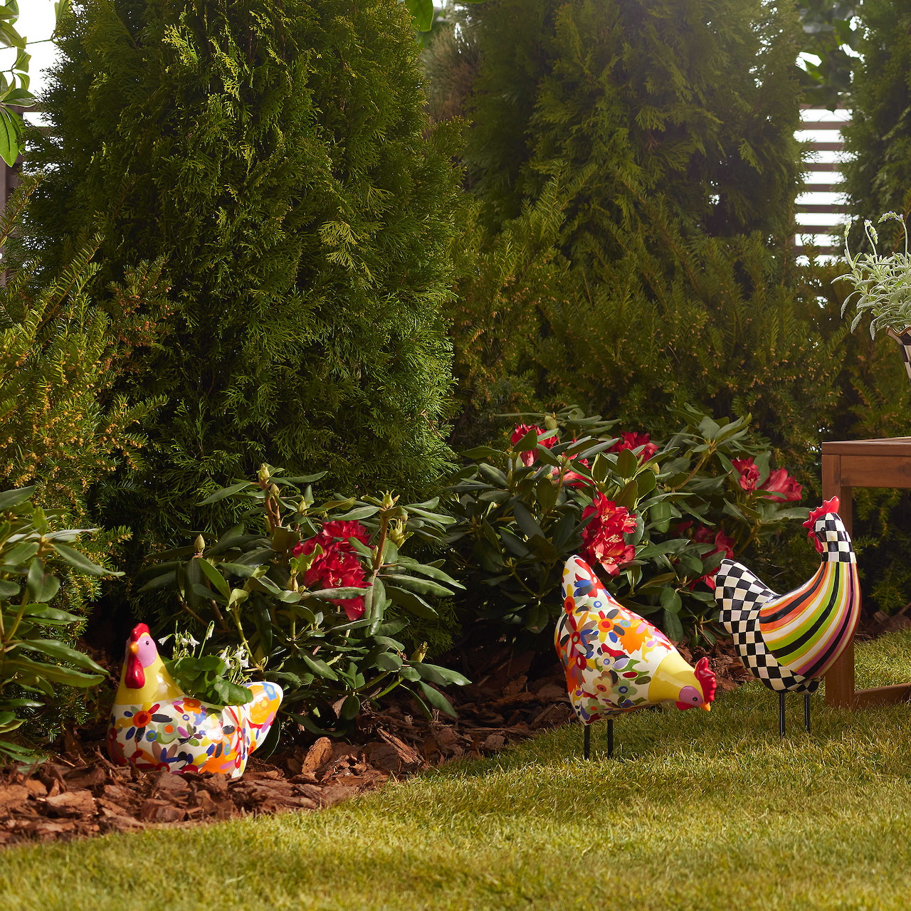 Two decorative ceramic chicken planters with red flowers on grass beside evergreen plants