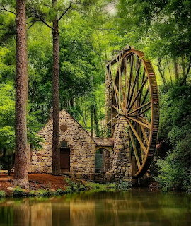 Stone mill ruins with large wooden water wheel surrounded by green trees and reflecting in pond