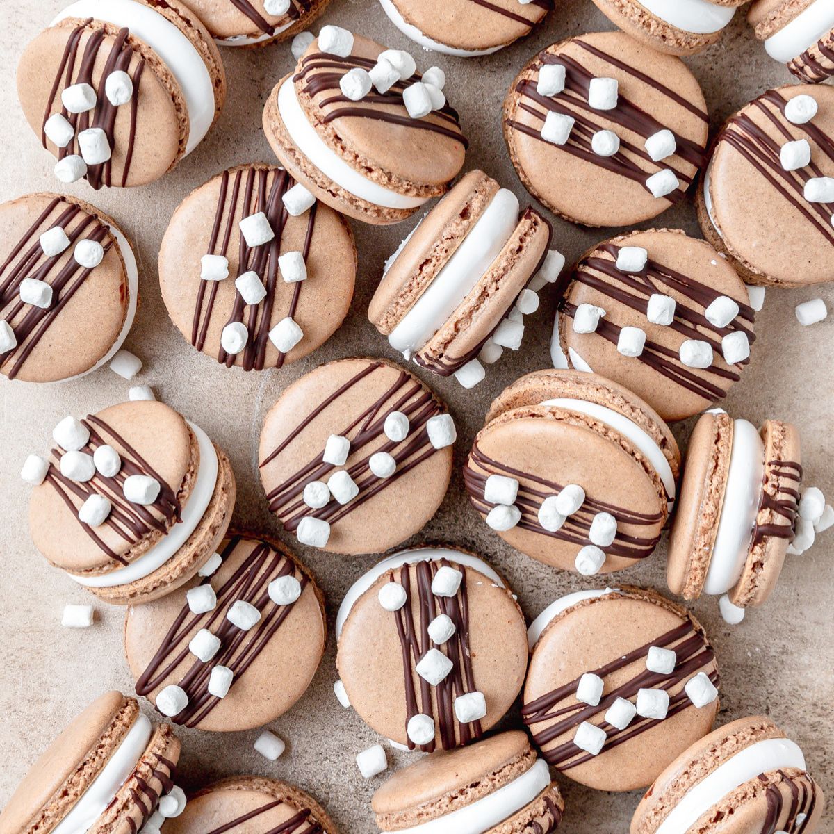 Chocolate macarons topped with icing drizzle and mini marshmallows on a light background