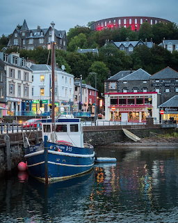 Blue fishing boat moored in Oban Bay with colorful waterfront buildings and hillside houses at dusk