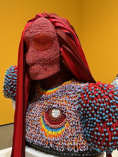Beaded sculpture of a figure with red fabric and colorful bead-covered surface in a gallery