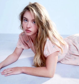 Sydney Sweeney lying on a light floor in a pale pink dress, looking at the camera