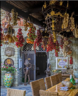 Rustic patio with hanging garlic, red peppers, wicker chairs, and a wooden table by a stone doorway