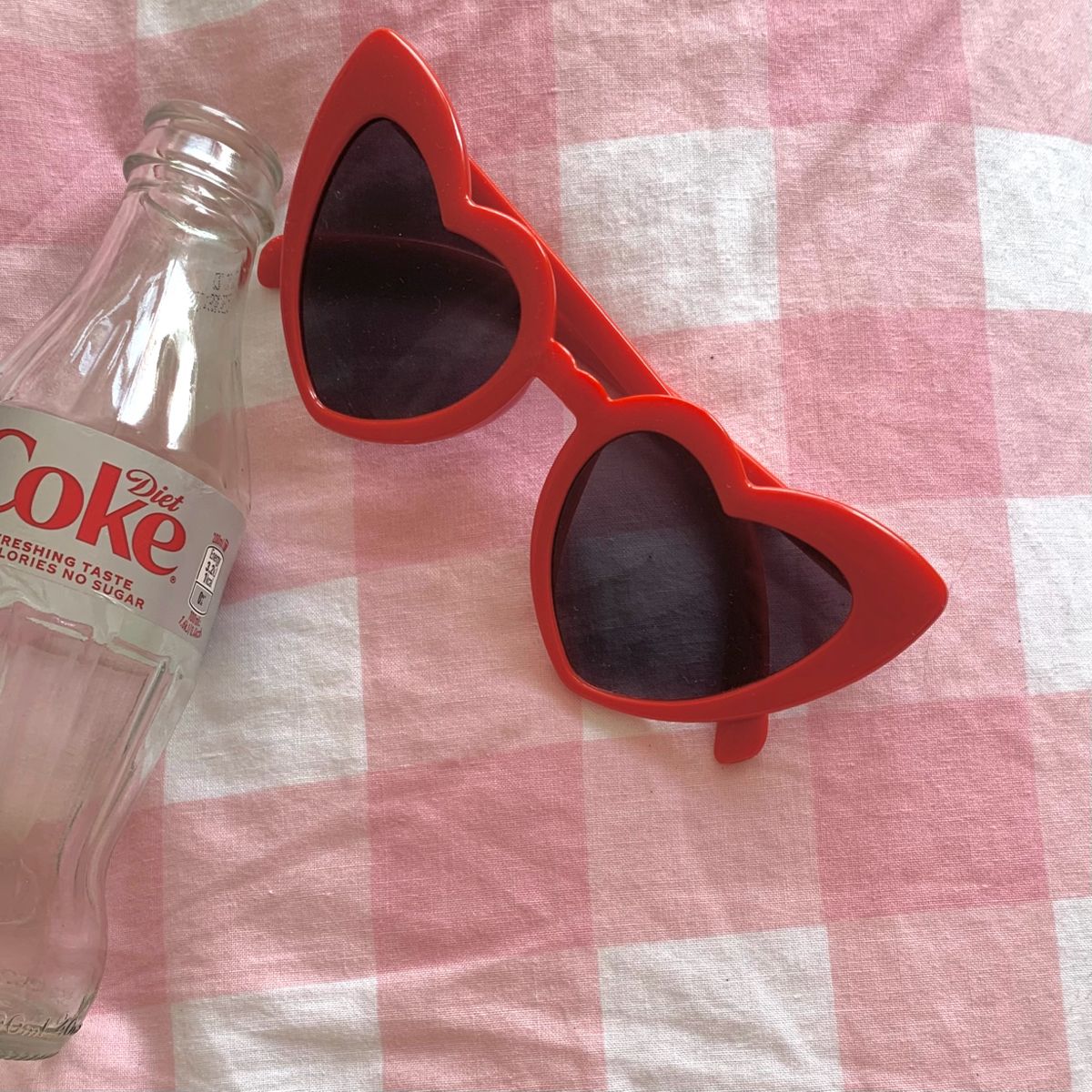 Red heart-shaped sunglasses next to a glass bottle on pink gingham fabric