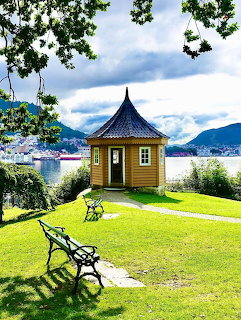 Yellow wooden pavilion on green lawn with wrought iron bench overlooking Bergen harbor and mountains