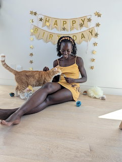 Woman in yellow dress sitting on floor petting orange cat with Happy Birthday banner on wall
