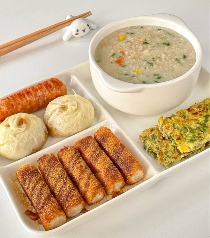 Korean meal tray with congee, fried tteok rice cakes, steamed buns, sausage, and vegetable pancake