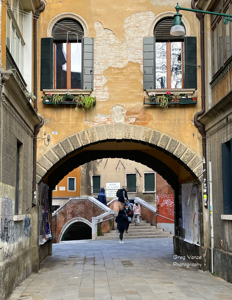 Weathered old city alley under a stone arch with a canal bridge and a person walking beyond