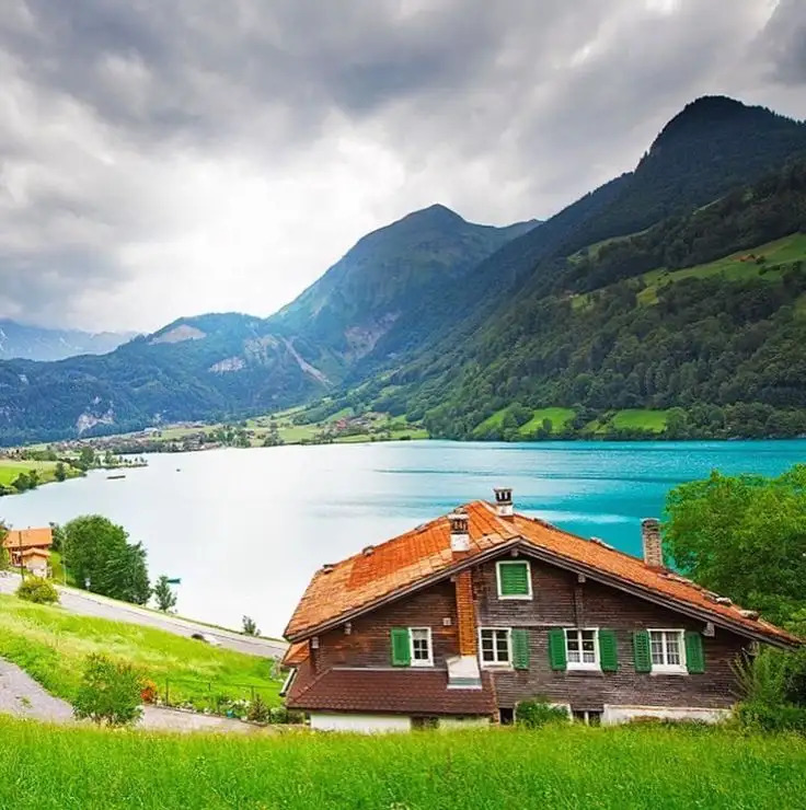 Wooden cabin above a turquoise lake with green mountains and cloudy sky
