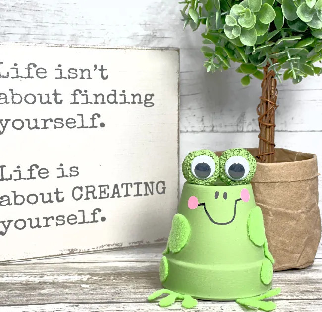 Green terracotta pot frog craft with googly eyes next to motivational sign and potted plant