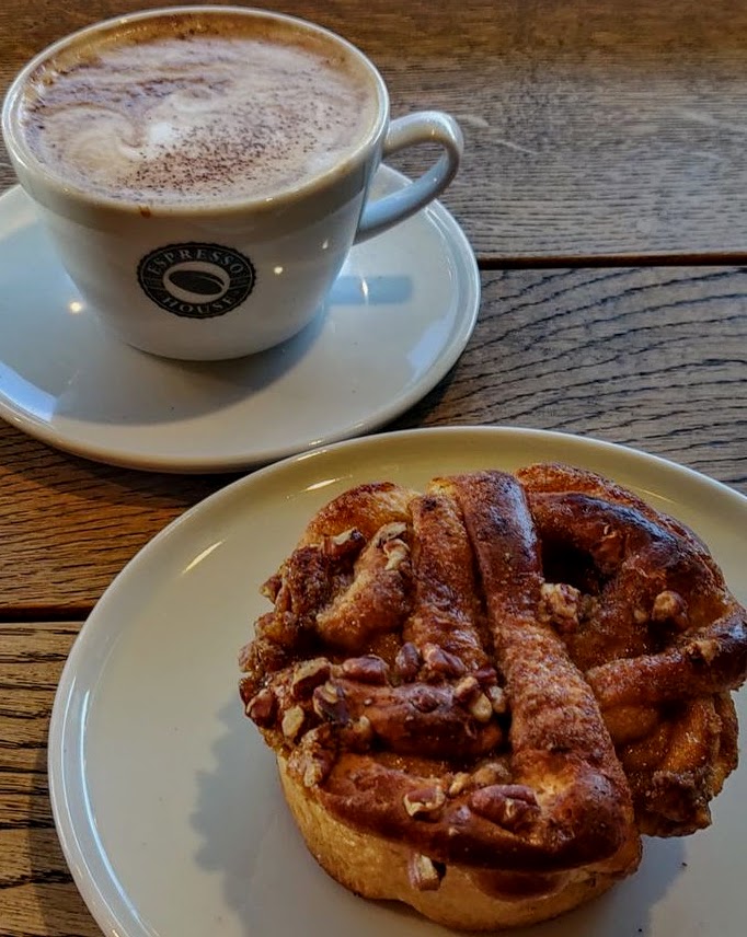 Cappuccino with foam and cinnamon roll with pecans on white plates at wooden table