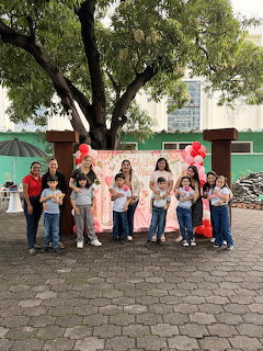 Group of children and adults posing in front of a pink backdrop with red balloons in a school courtyard