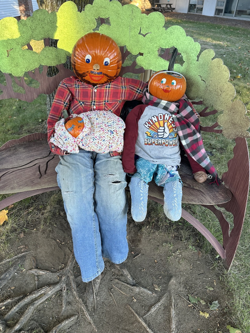 Two pumpkin-headed scarecrows sitting on a bench dressed in casual clothing with a small pumpkin baby