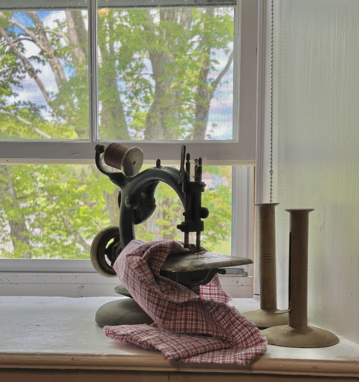 Antique sewing machine with plaid fabric on a windowsill beside brass candlestick holders, green trees in background