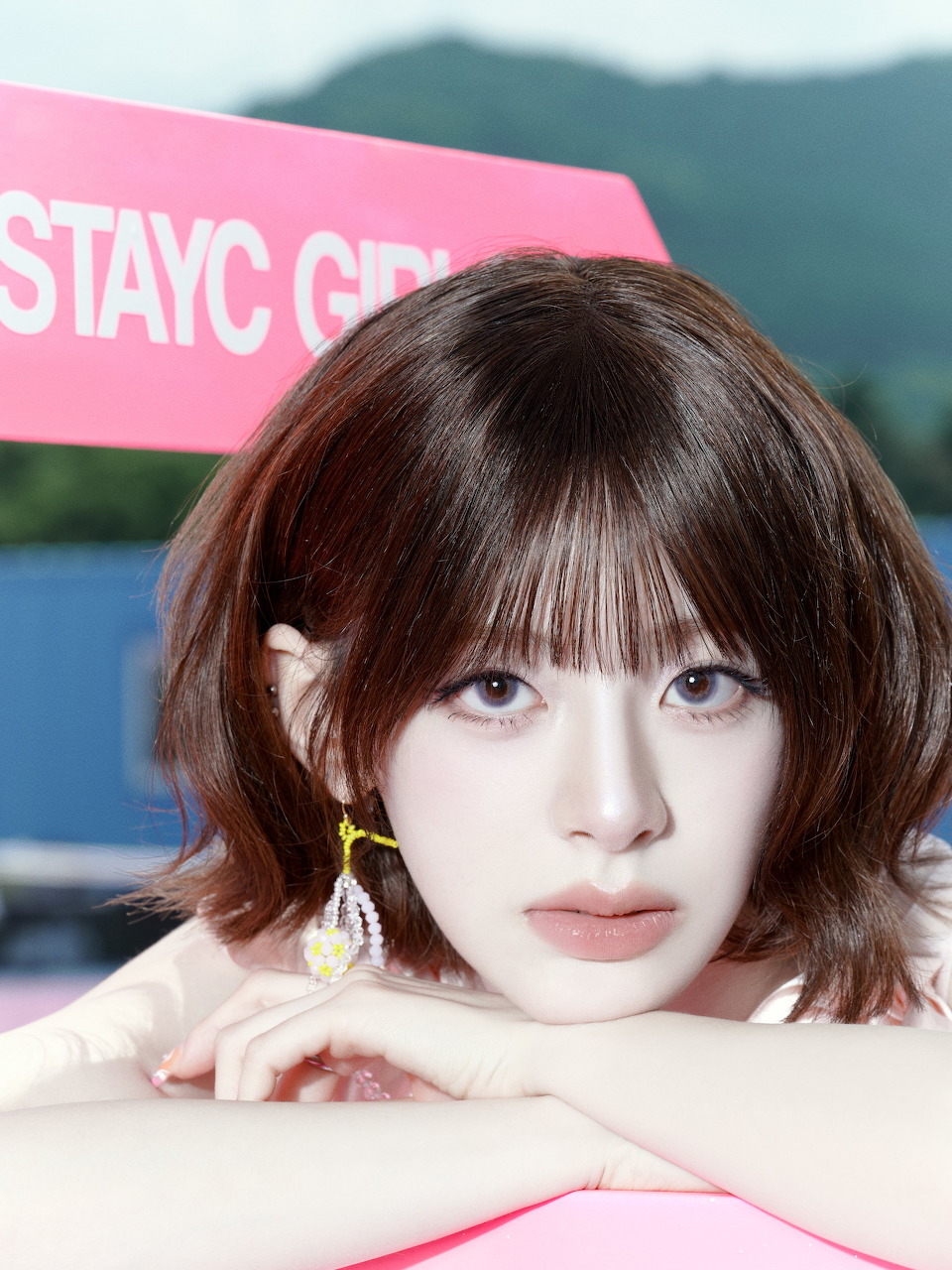Portrait of young woman with short brown hair and bangs wearing white, resting chin on hands against pink and blue background