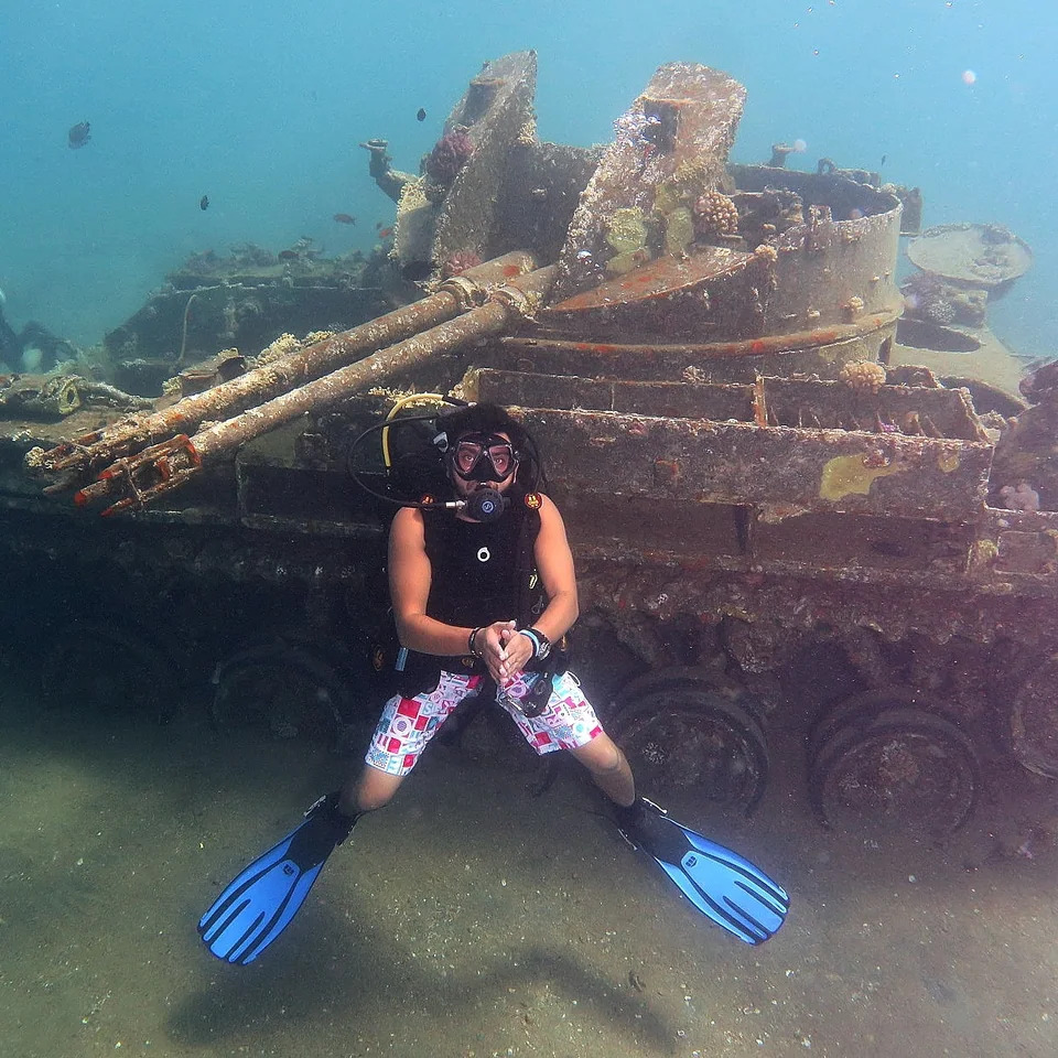 Scuba diver posing in front of sunken military vehicle covered in coral underwater
