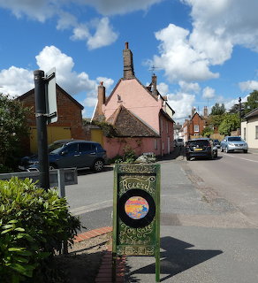 Village street with parked cars, a pink building, and a sign beside the road
