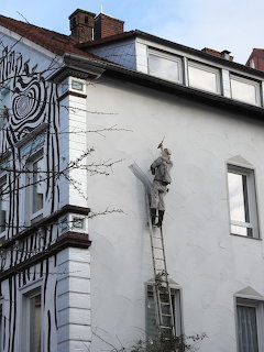 Sculpture of a figure climbing a ladder on a white European building facade with black mural art