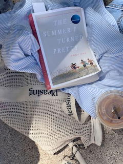 Book titled The Summer I Turned Pretty on blue striped cloth with iced coffee, sunglasses, and mesh bag