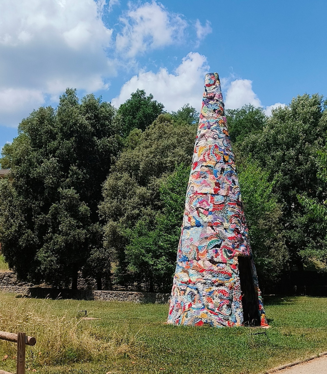 Tall cone-shaped mosaic sculpture with vibrant colors standing in a grassy park with trees in the background.