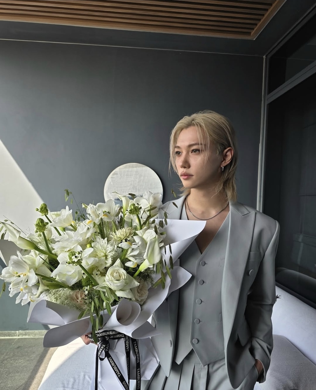 Blonde woman in a gray suit standing beside a bouquet of white flowers indoors