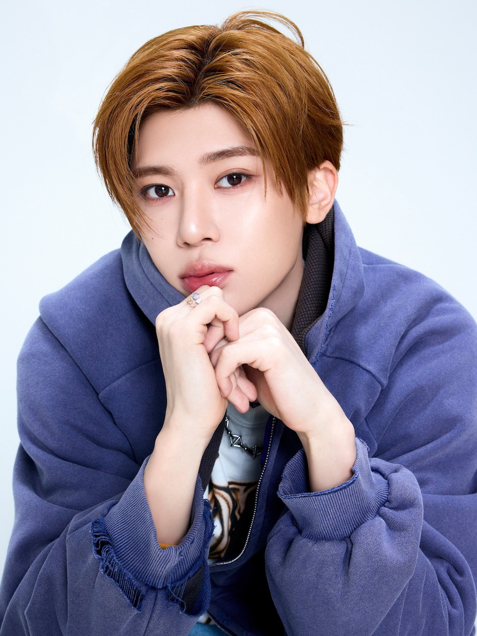 Young East Asian man with auburn hair in a blue hoodie, hands near chin, light background
