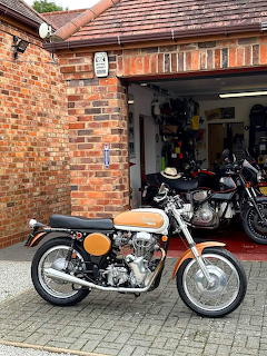 Orange vintage motorcycle parked on driveway in front of brick garage with black motorcycle inside