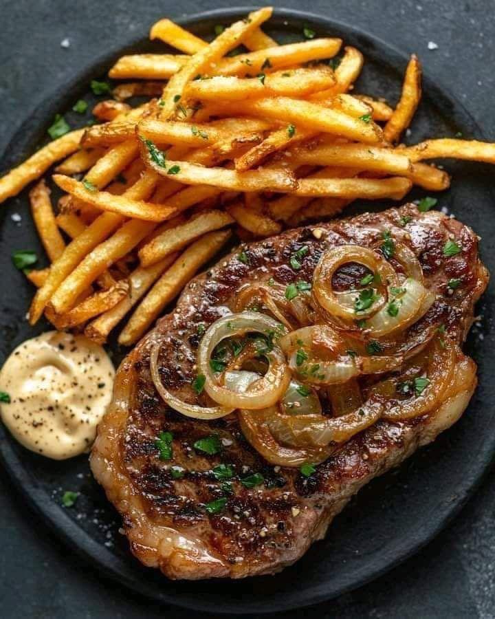 Grilled steak with caramelized onions and herbs alongside crispy french fries on a dark plate