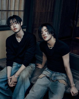 Two young men in black tops and jeans posing together in a dark indoor setting