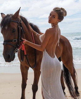 Woman in white dress standing next to brown horse on beach with ocean and sky