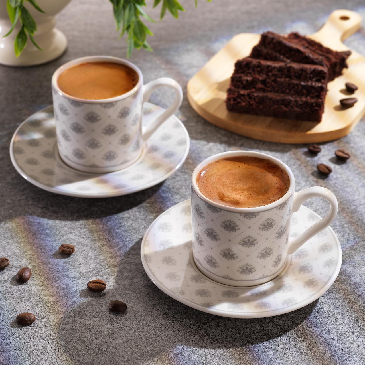 Two white patterned coffee cups with espresso and a slice of chocolate cake on a wooden plate with scattered coffee beans