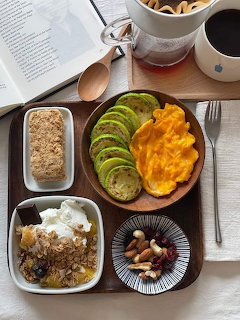 Wooden breakfast tray with fruit, eggs, granola, yogurt, nuts, coffee and open book on white surface