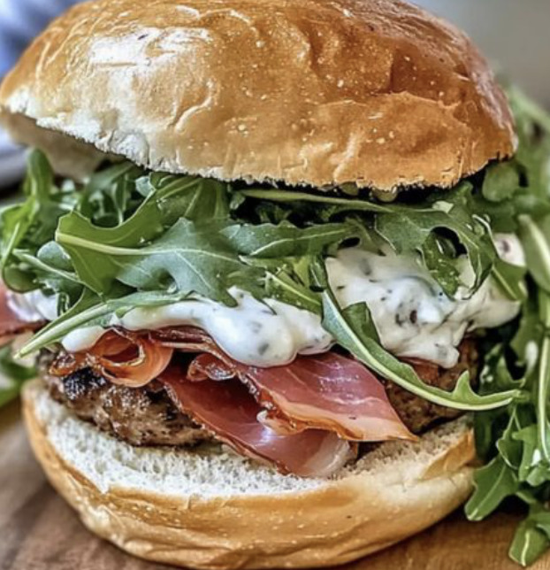 Close-up of a burger topped with arugula, prosciutto, creamy sauce, and a shiny bun