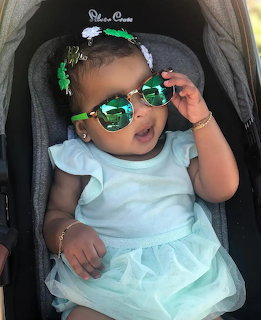 Baby wearing green mirrored sunglasses and floral headband sitting in a stroller with a tu dress