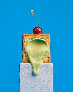 Square pastry with dripping green cream and a red cherry on a pedestal against a blue backdrop
