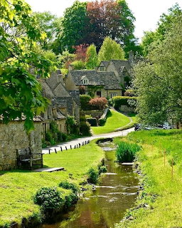 Historic stone cottages beside a gentle stream in an English village, framed by green lawns and trees