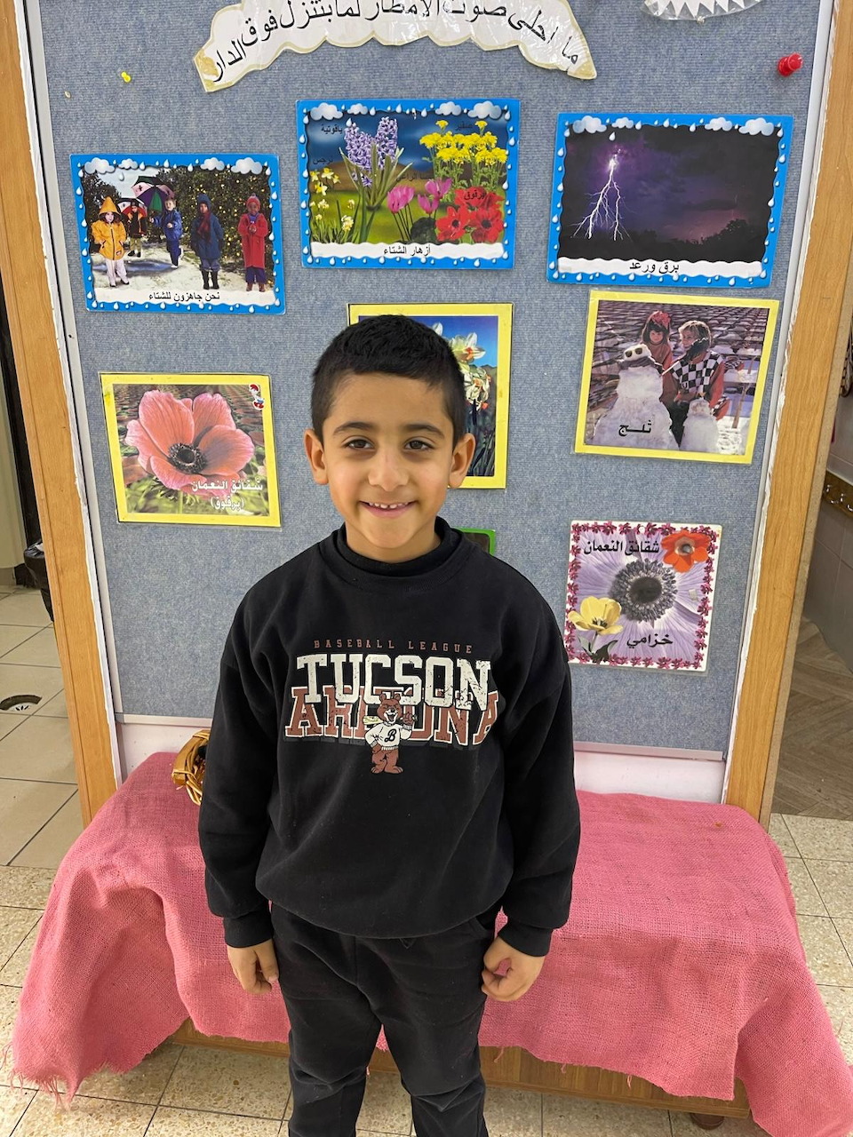 Young boy in black Tucson sweatshirt smiling in front of a colorful school bulletin board with artwork