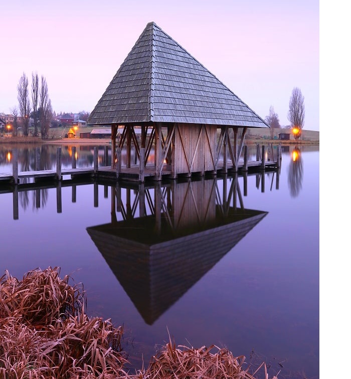 Wooden pyramid pavilion reflected in calm pond water at dusk with purple sky