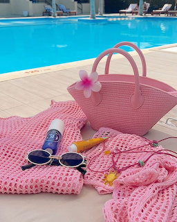 Pink woven bag and accessories on pink towel beside blue swimming pool