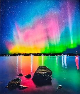 Colorful northern lights reflecting on calm lake water with rocks in foreground