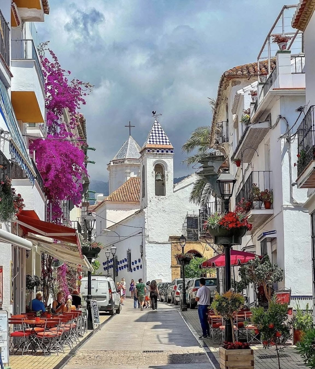 Narrow pedestrian street in Marbella old town with white buildings, purple flowers, and church bell tower