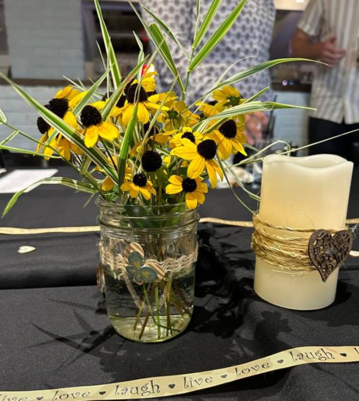 Yellow black-eyed Susan flowers in a mason jar next to a pillar candle on a dark table with a decorative ribbon