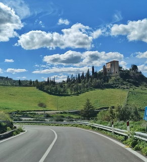 Winding road leading to an old palazzo on a green hillside with cypress trees and blue sky