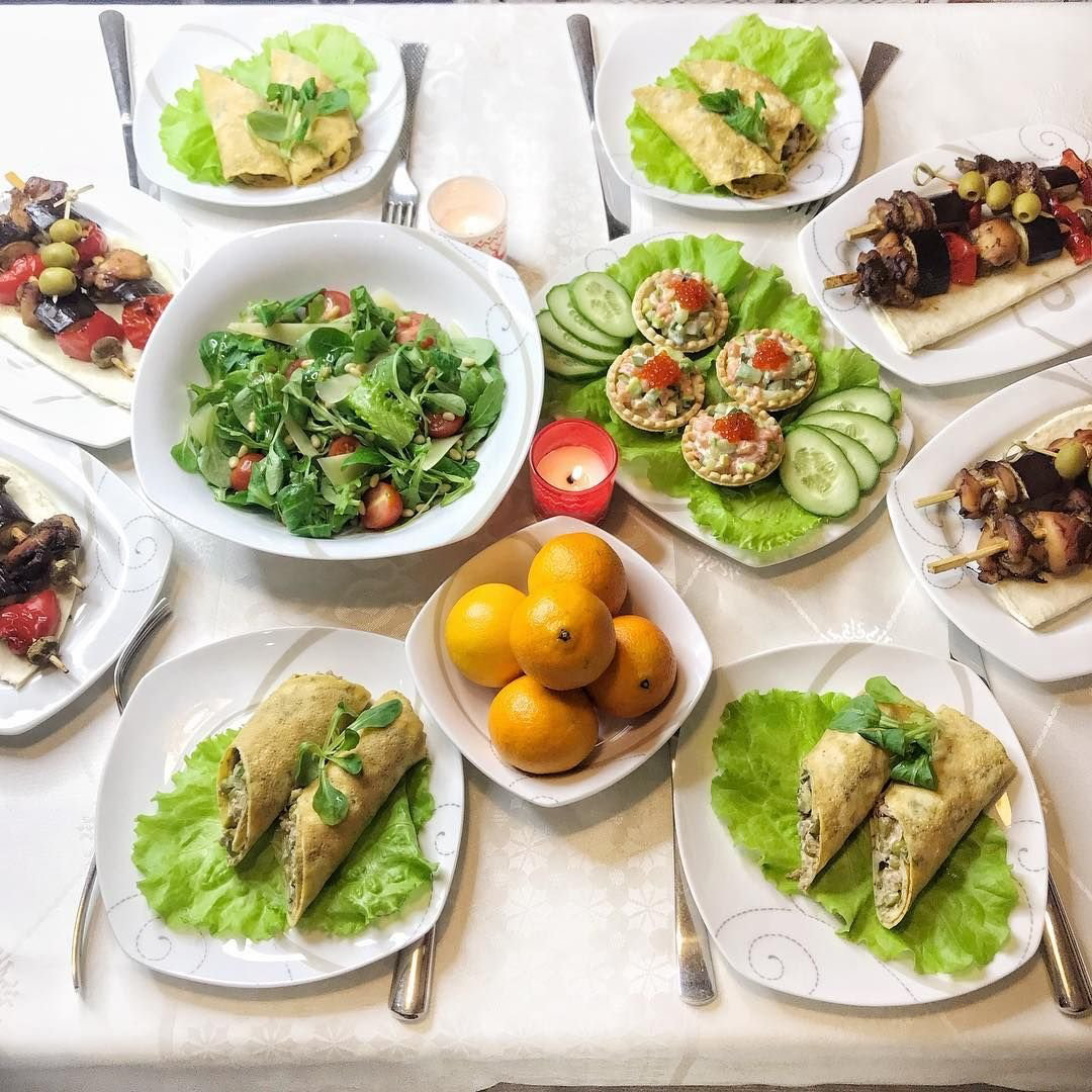 Overhead view of multiple white plates with healthy meals, salads, wraps, and fresh fruit on a light surface